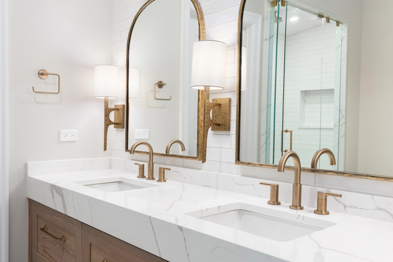 A bathroom detail with gold faucets, light fixtures, and mirrors, a wood cabinet, large marble slab countertop, and a subway tile shower